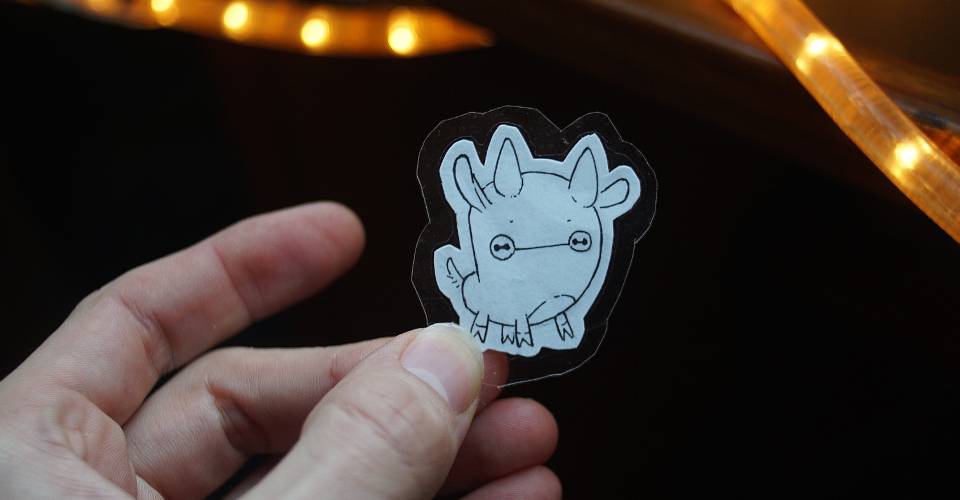A photo of a hand holding a cut-out of a hand-drawn version of the uxn logo, a little ox-like creature